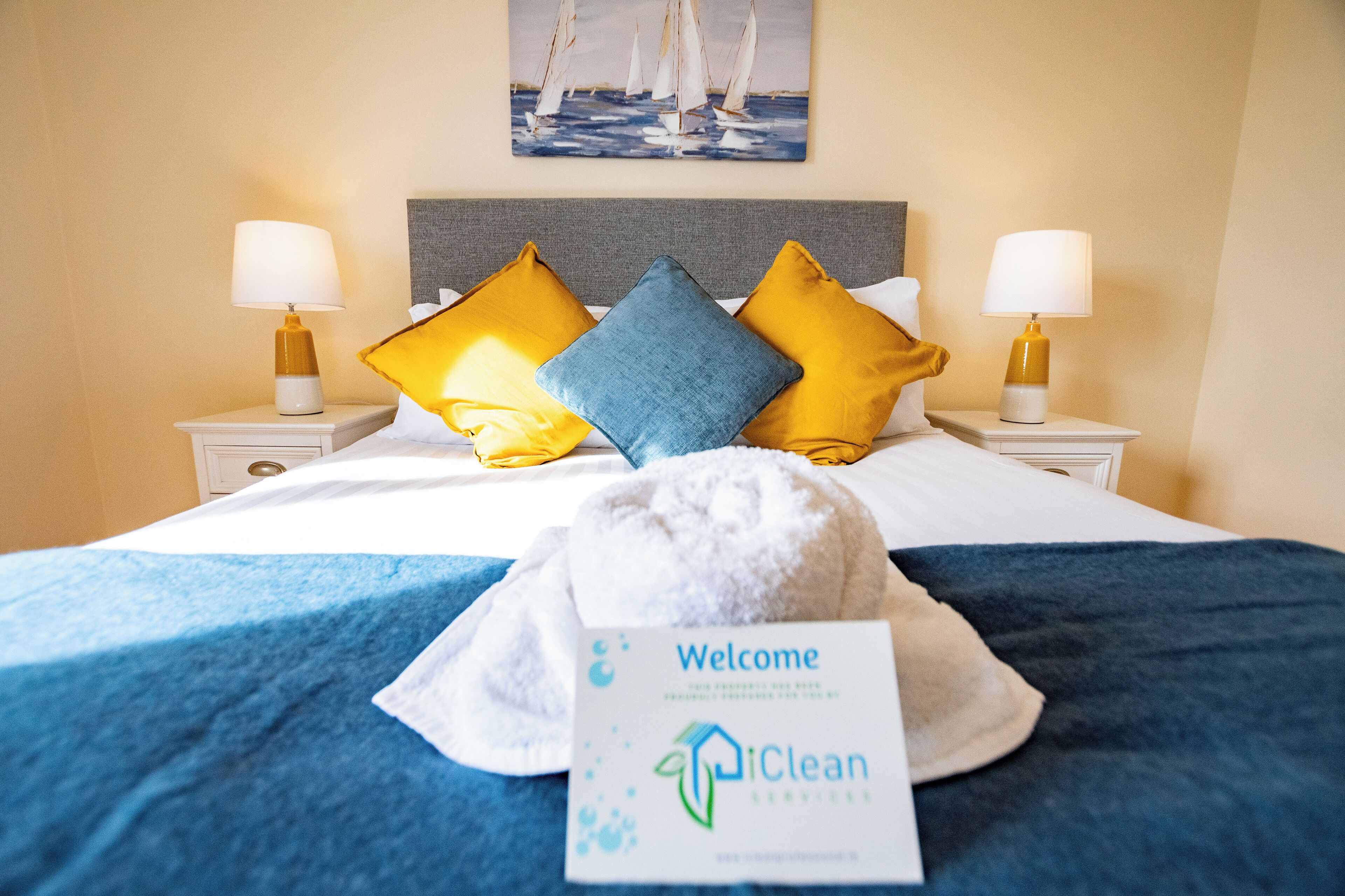 A neatly made bed with a blue bedspread, two pillows in yellow and blue, and a towel on the bed. There is a welcome sign on the bed and a painting of sailboats on the wall.