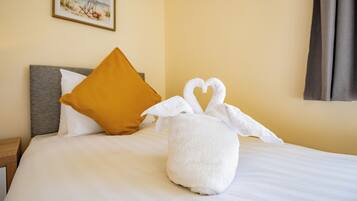 A neatly made bed with a white pillow, a mustard-colored pillow, and a white towel shaped like a bunny.