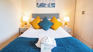 Ein ordentlich bezogenes Bett mit einer Kissenanordnung in Blau und Gelb, ein Willkommensschild und ein Gemälde mit Segelbooten an der Wand.