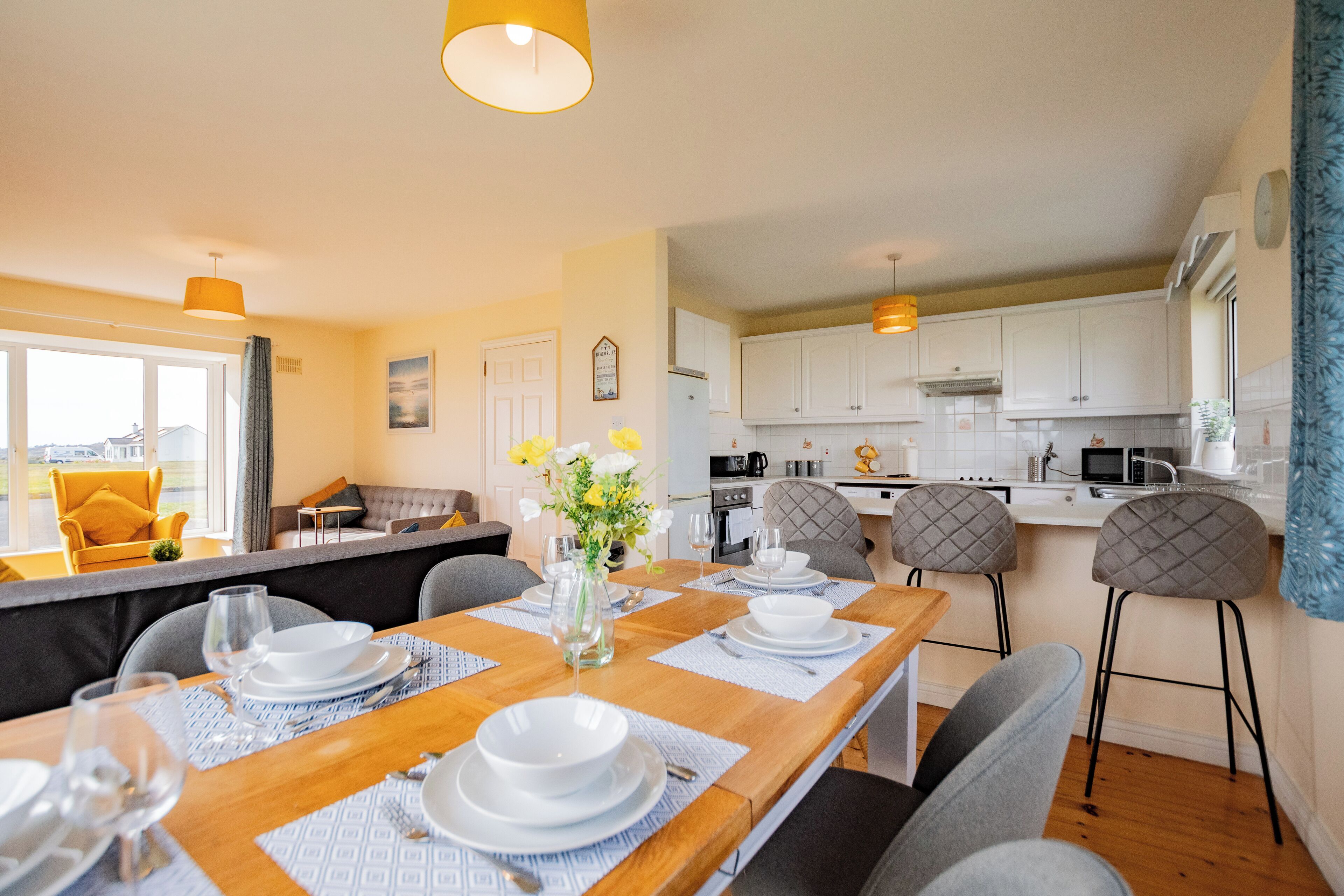 Una cocina moderna con armarios blancos, un comedor con mesa de madera dispuesta para cuatro comensales, y vistas del área de estar con un sofá y una ventana.