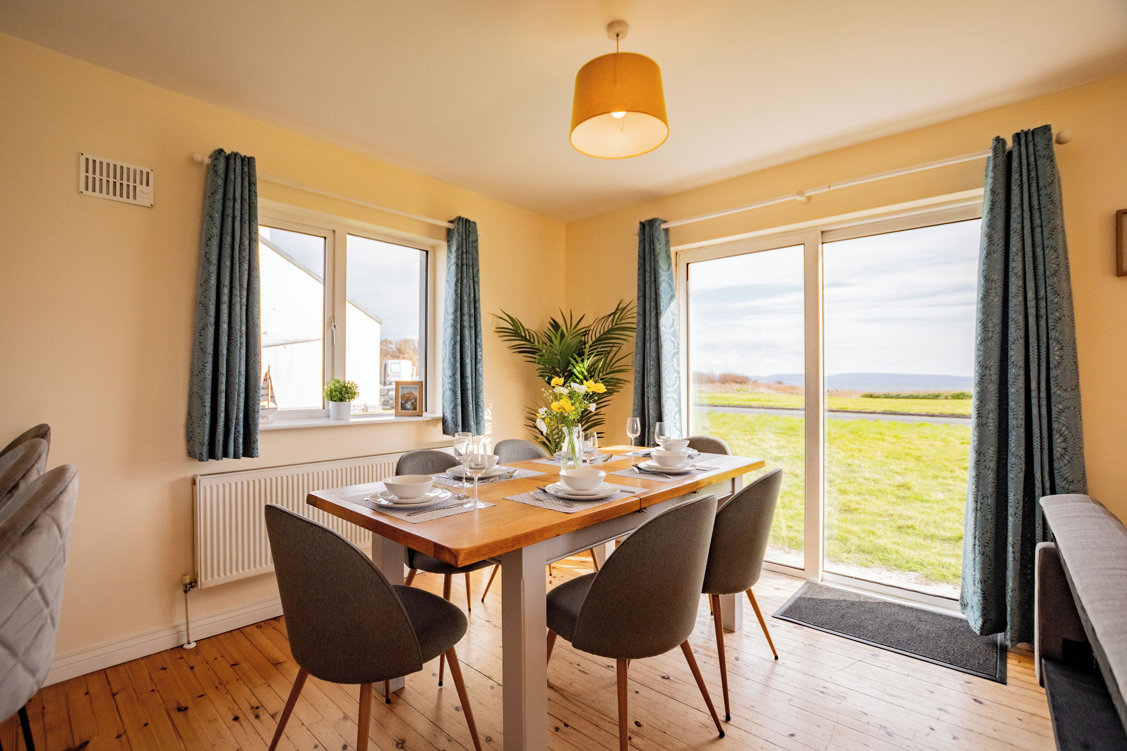 A dining area with a wooden table set for four, surrounded by chairs, a potted plant, and a view of the outdoors.