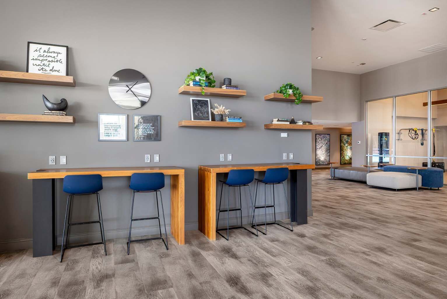 A modern kitchen with bar stools, wooden shelves with plants, a clock, and framed quotes on the wall.