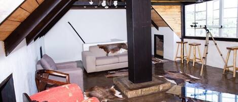 A loft-style room with a sloped ceiling, wooden beams, a sofa, a red chair, a table, and a large window.
