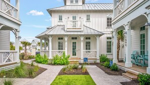 Une grande maison blanche à deux étages, dotée d’un porche, d’un petit jardin et surplombant un ciel bleu dégagé.