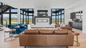 A modern living room with a large sofa, a coffee table, and a fireplace. There is a kitchen area with cabinets and a countertop. A staircase is visible in the background. - Hot Tub, 23 Mi to Grand Targhee: Luxe Retreat (Tetonia)