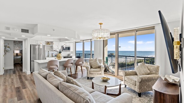 Un vaste espace de vie comprenant un grand canapé, une table basse et deux fauteuils. On aperçoit une cuisine au fond de la pièce, et de grandes fenêtres offrent une vue sur l’océan.