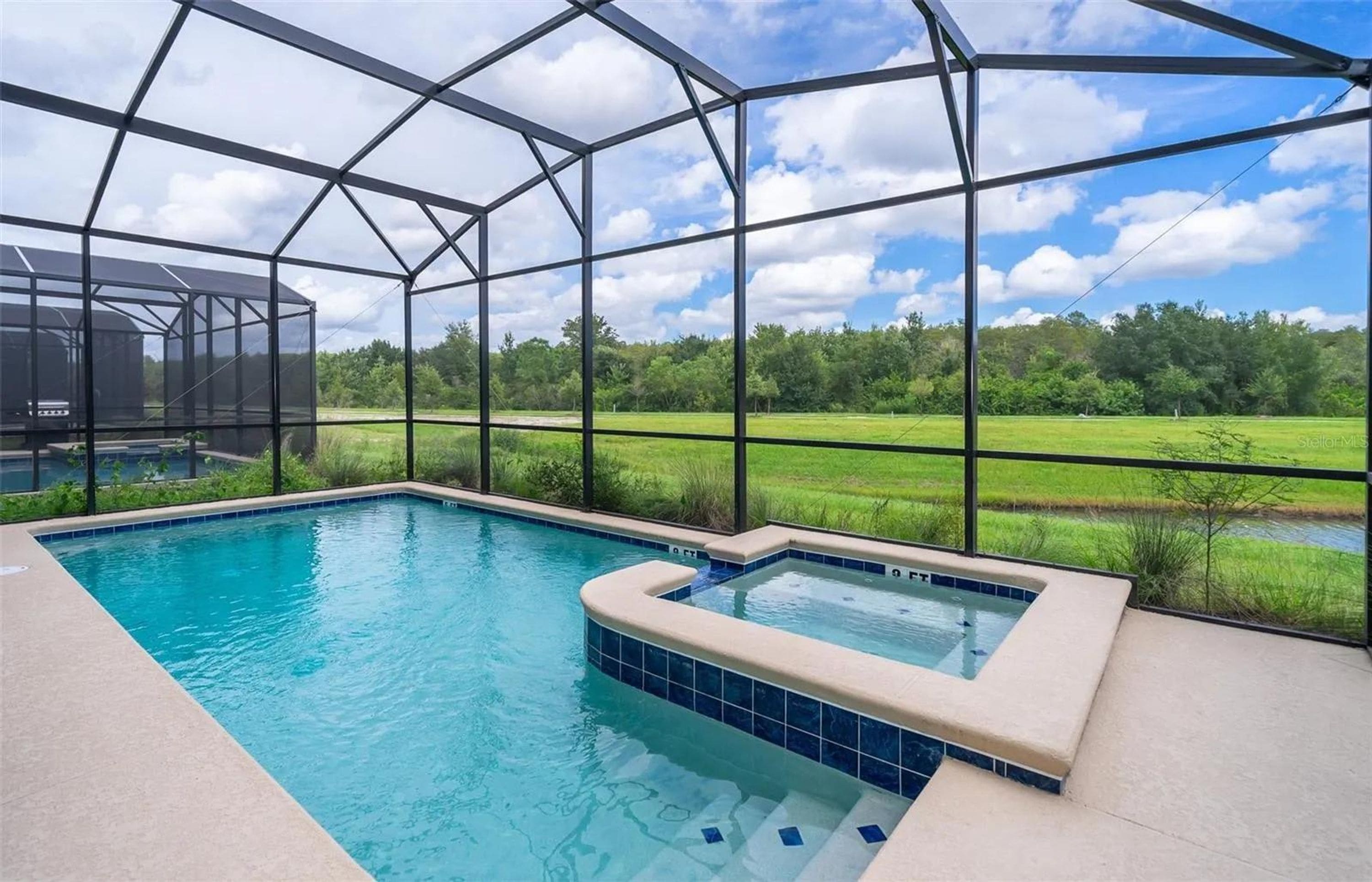 Une piscine avec un jacuzzi, entourée d’une structure en verre offrant une vue sur un champ verdoyant et des arbres.