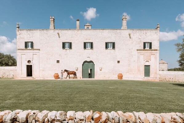 A large, white stone building with a green door and windows, a horse in front, and a stone wall in the foreground. - Masseria Lamacoppa Exclusive Estate Near Ostuni (Ostuni)