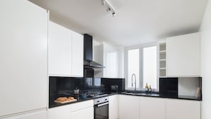 Une cuisine moderne avec des meubles blancs, un plan de travail noir, une hotte en acier inoxydable et une fenêtre qui laisse entrer la lumière naturelle.
