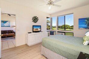 A bedroom with a bed, a television on a cabinet, a large window with a view, and a ceiling fan.