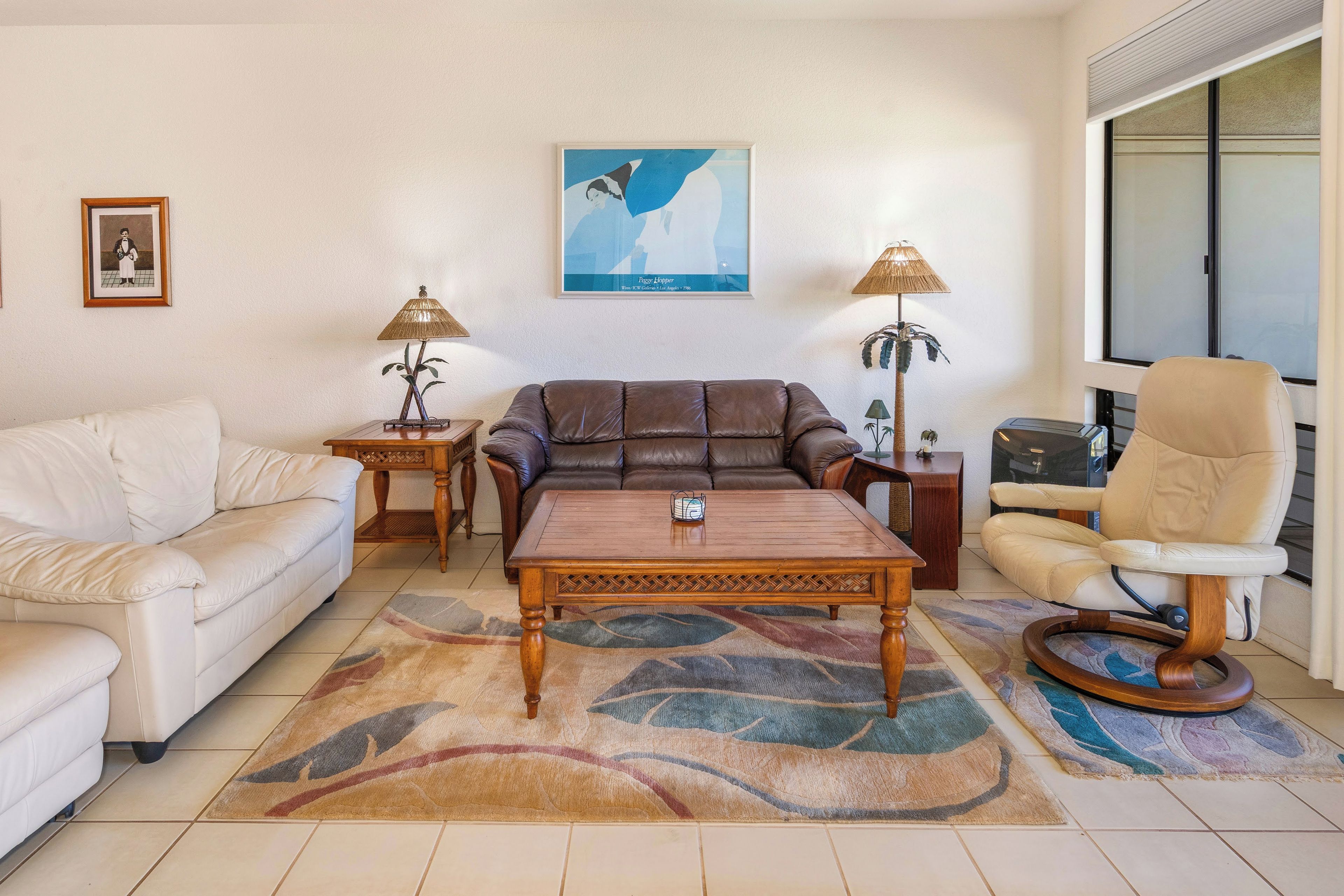 Una sala de estar con un sofá de cuero marrón, un sofá blanco, una mesa de centro de madera, dos mesas auxiliares con lámparas y un sillón color beige.