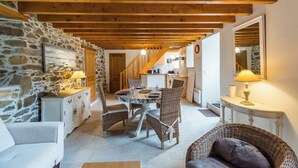 Un interior acogedor con pared de piedra, vigas de madera, una zona de comedor con sillas de mimbre y una cocina al fondo.