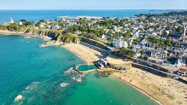 Vista aérea de un pueblo costero con playa, casas y un faro.