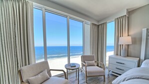 Une chambre d’hôtel dotée d’une grande fenêtre donnant sur la plage, avec un lit, une chaise, une petite table et une table de chevet avec une lampe.