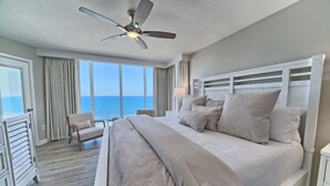 Une chambre avec un grand lit, un ventilateur de plafond, une chaise et une vue sur l’océan.