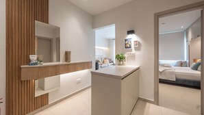 Ein moderner, gut beleuchteter Flur mit einer Wand in Holzoptik, einer zentralen Kücheninsel mit Spüle und Blick in ein Schlafzimmer mit Bett und Schlafsofa.