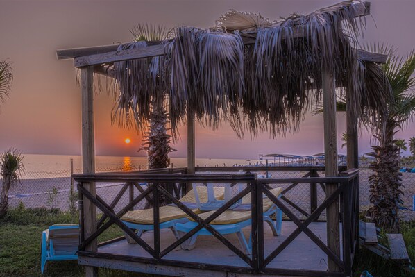 A wooden gazebo with a thatched roof, blue lounge chairs, and a view of the sunset over the ocean. - Elysium Elite Hotel&Spa (Manavgat)