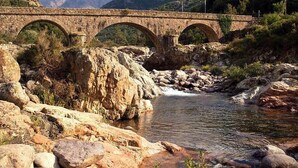 Un puente de piedra con tres arcos sobre un río, rodeado de terreno rocoso y montañas.