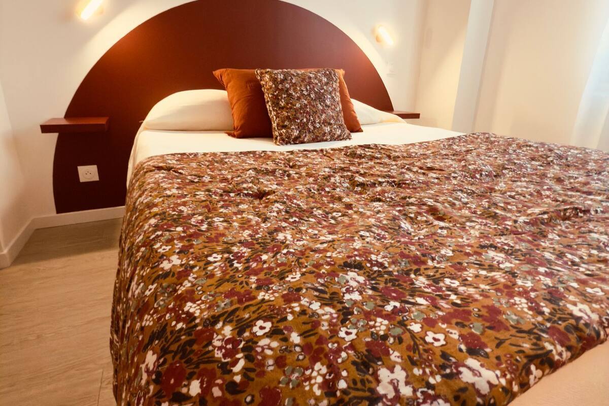 Een net opgemaakt bed met een bloemenbedekking, een houten hoofdbord en een nachtkastje met een lamp.