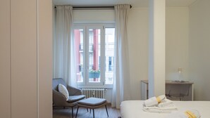 A hotel room with a bed, a chair, a small table, and a window with curtains. - Brand new 2 BR With Balcony in Sempione (Milan)