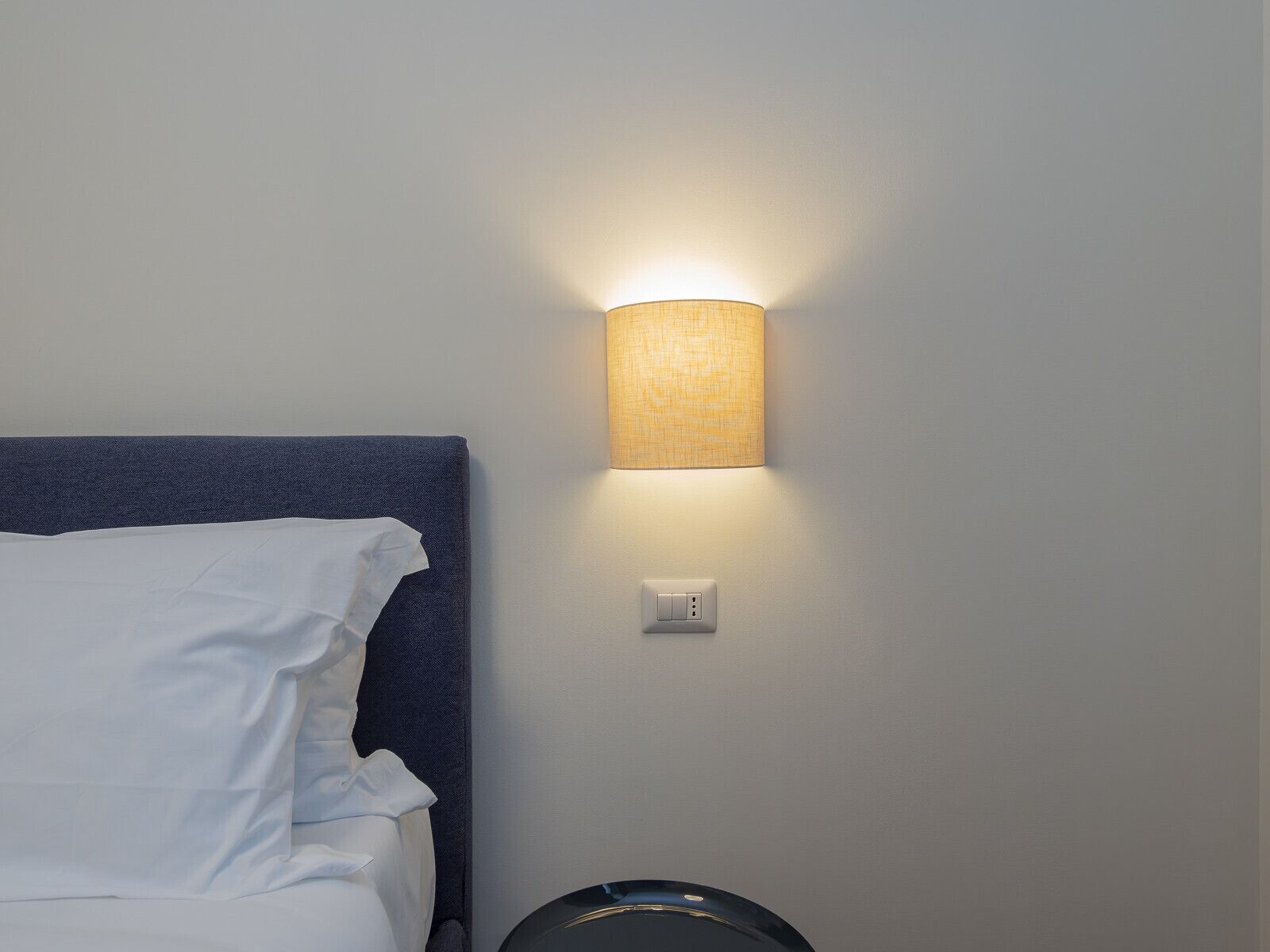 Un lit bien fait, avec du linge de lit blanc et une tête de lit bleue, une lampe fixée au mur et une prise de courant.