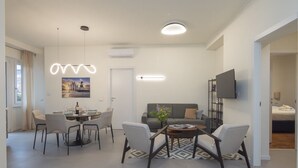 A modern living room with a dining area, grey sofas, a flat-screen TV, and a large window. - Brand new 2 BR With Balcony in Sempione (Milan)