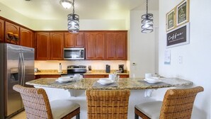 A kitchen with wooden cabinets, a stainless steel refrigerator, a microwave, and a granite countertop. There are wicker chairs around the island, and two pendant lights hang above. - Pool View Penthouse 3 Bedroom Condo (Orlando)