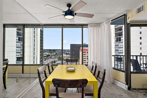 Un balcon avec une table et des chaises pour prendre les repas, un ventilateur de plafond et une vue sur le paysage urbain, visible à travers de grandes fenêtres.