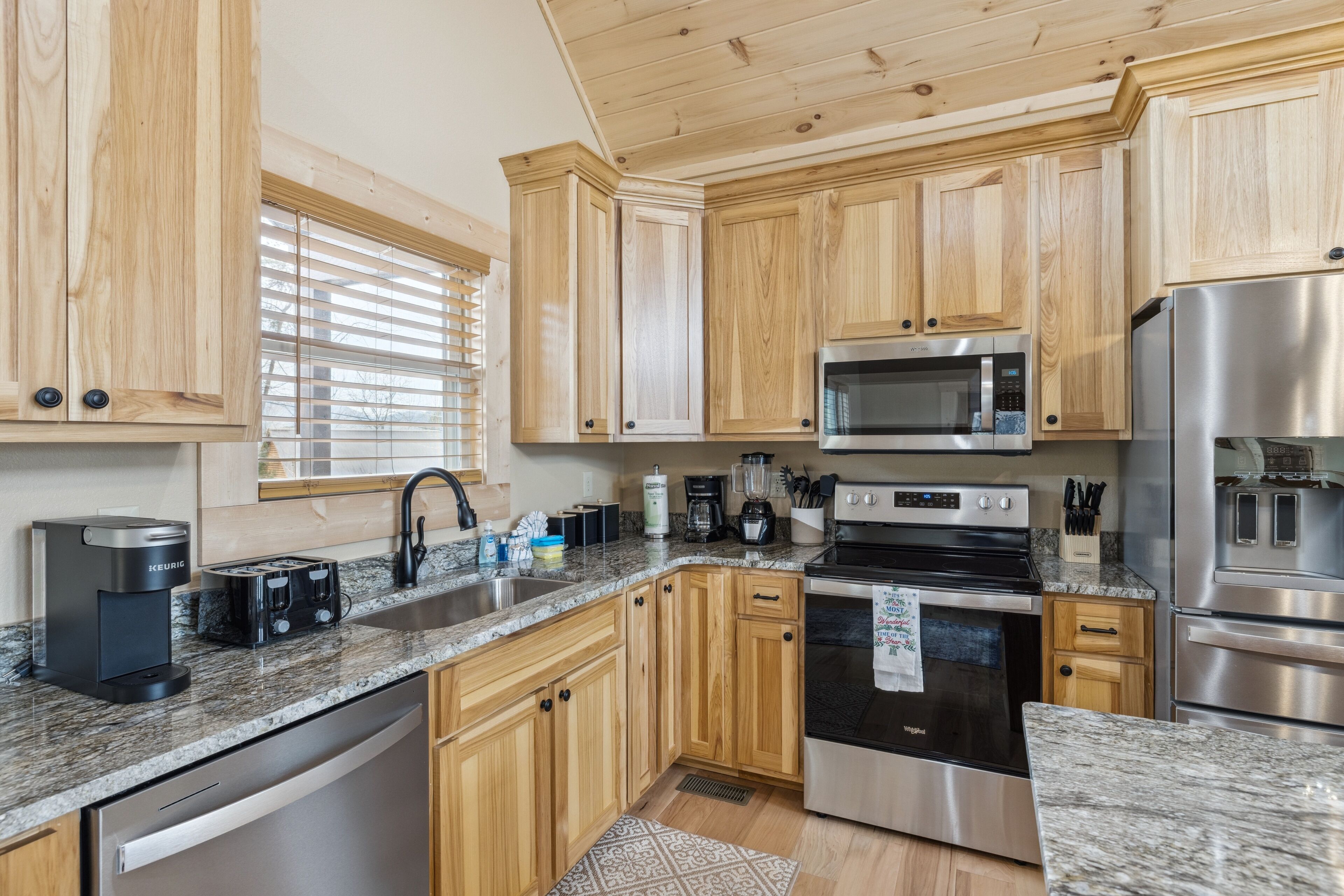 A modern kitchen with wooden cabinets, stainless steel appliances, granite countertops, and a window with blinds.