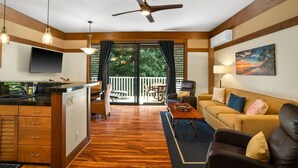 A modern living room with a wooden floor, a ceiling fan, a flat-screen TV, a sofa, a coffee table, and a dining area with a table and chairs. - Kauai Kiahuna Plantation #425 1 Bedroom Condo (Koloa)