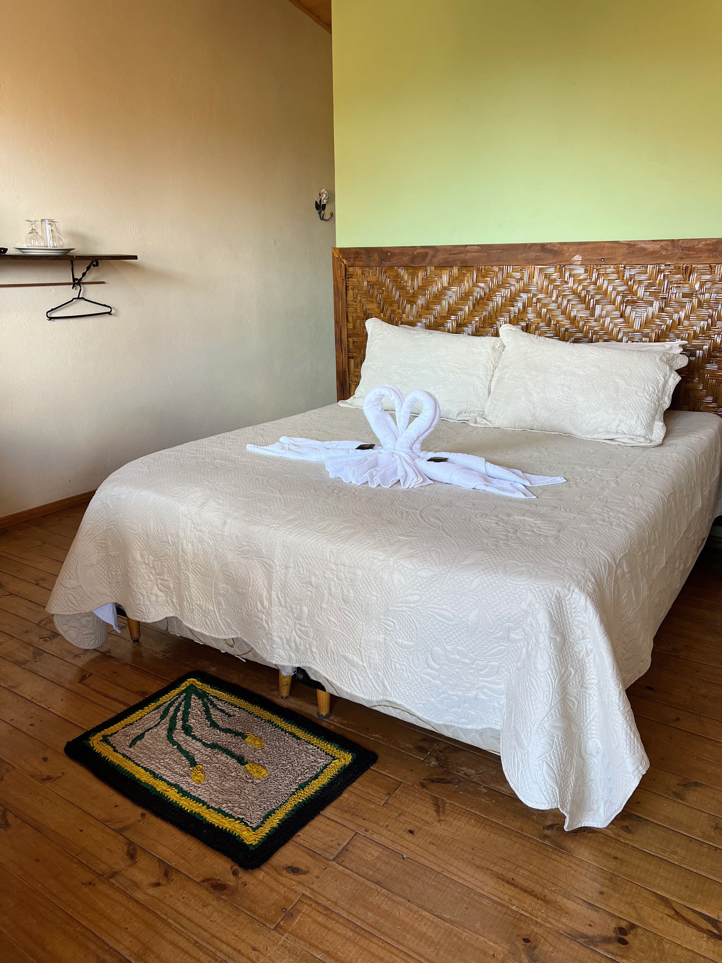 A neatly made bed with a white bedspread and two pillows, a wooden headboard with a woven pattern, and a small hanging shelf with a glass container and a hanger.