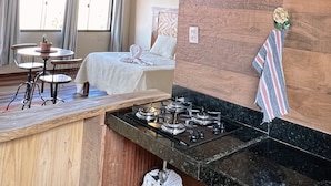 A room with a wooden ceiling, a bed with white linens, a dining table with chairs, and a kitchen area with a stove.