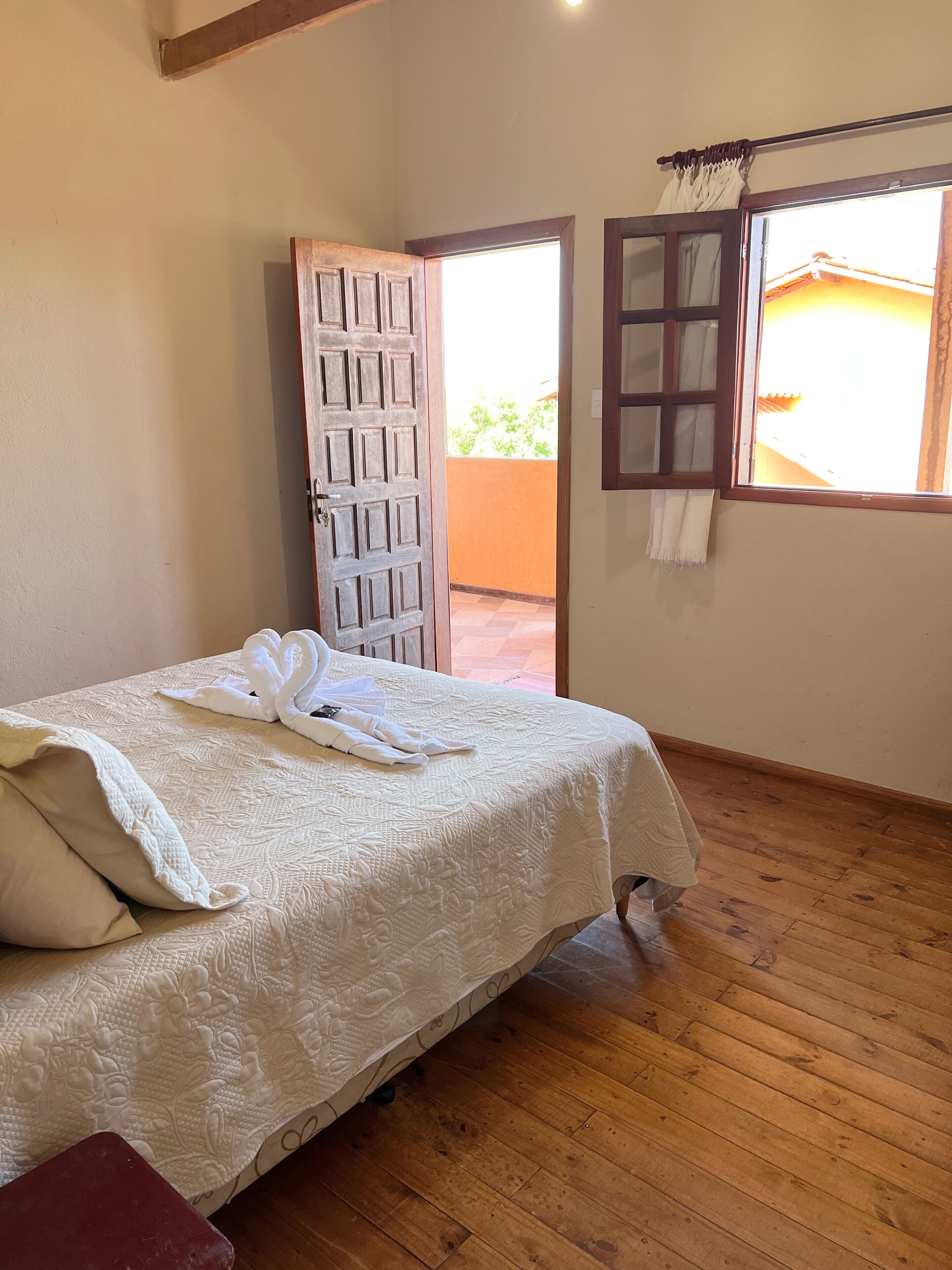 A hotel room with a wooden floor, a bed with a white bedspread, and a window with curtains.