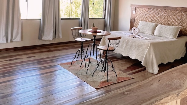 A room with a wooden ceiling, a bed with a woven headboard, a round table with chairs, and a window with curtains.