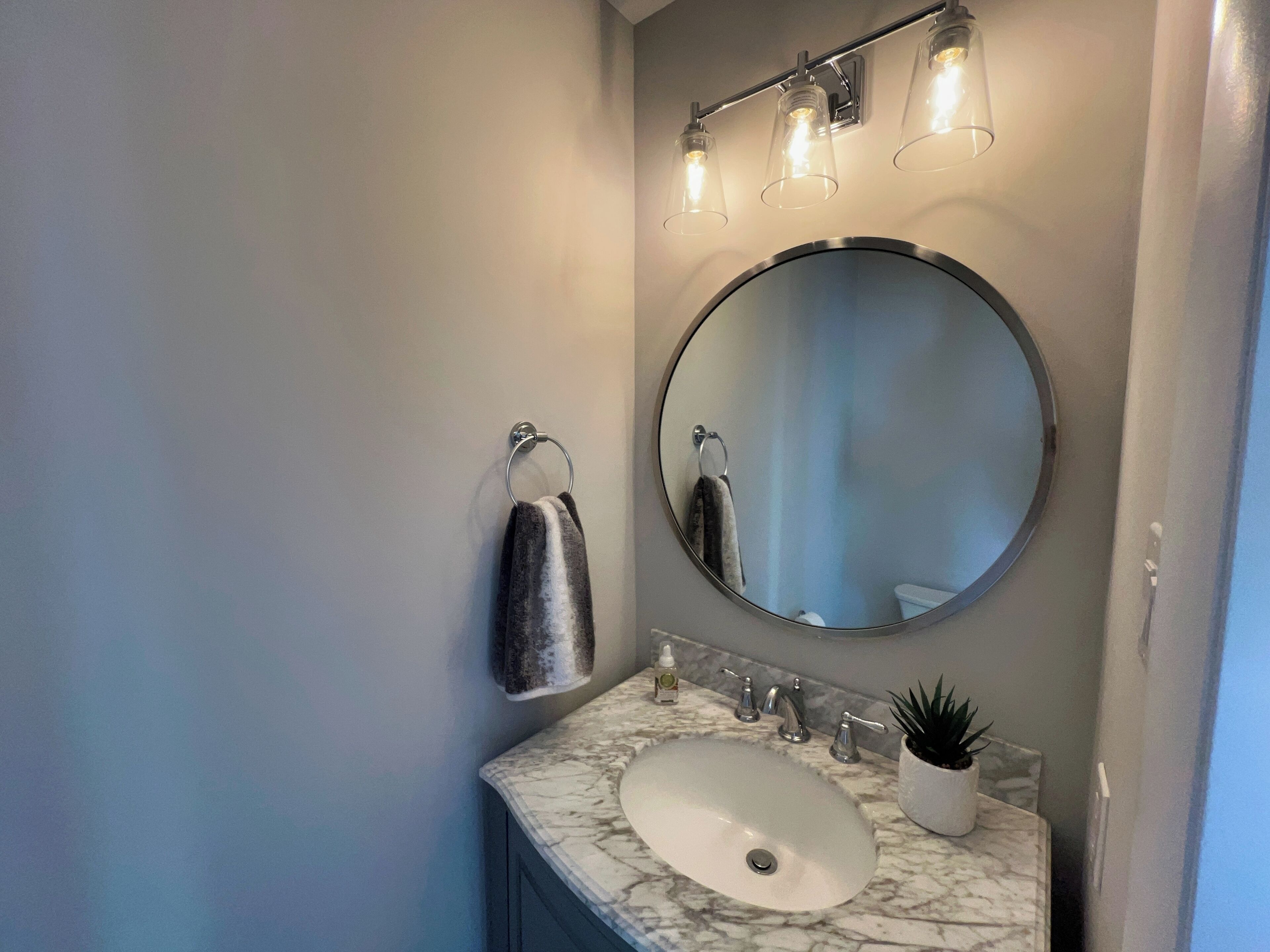 A bathroom with a round mirror, a white sink, and a marble countertop. There are three light fixtures above the mirror and a towel rack on the wall.