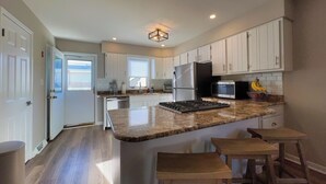 A modern kitchen with white cabinets, a stainless steel refrigerator, a microwave, and a gas stove. There are wooden stools at the island and a window providing natural light.