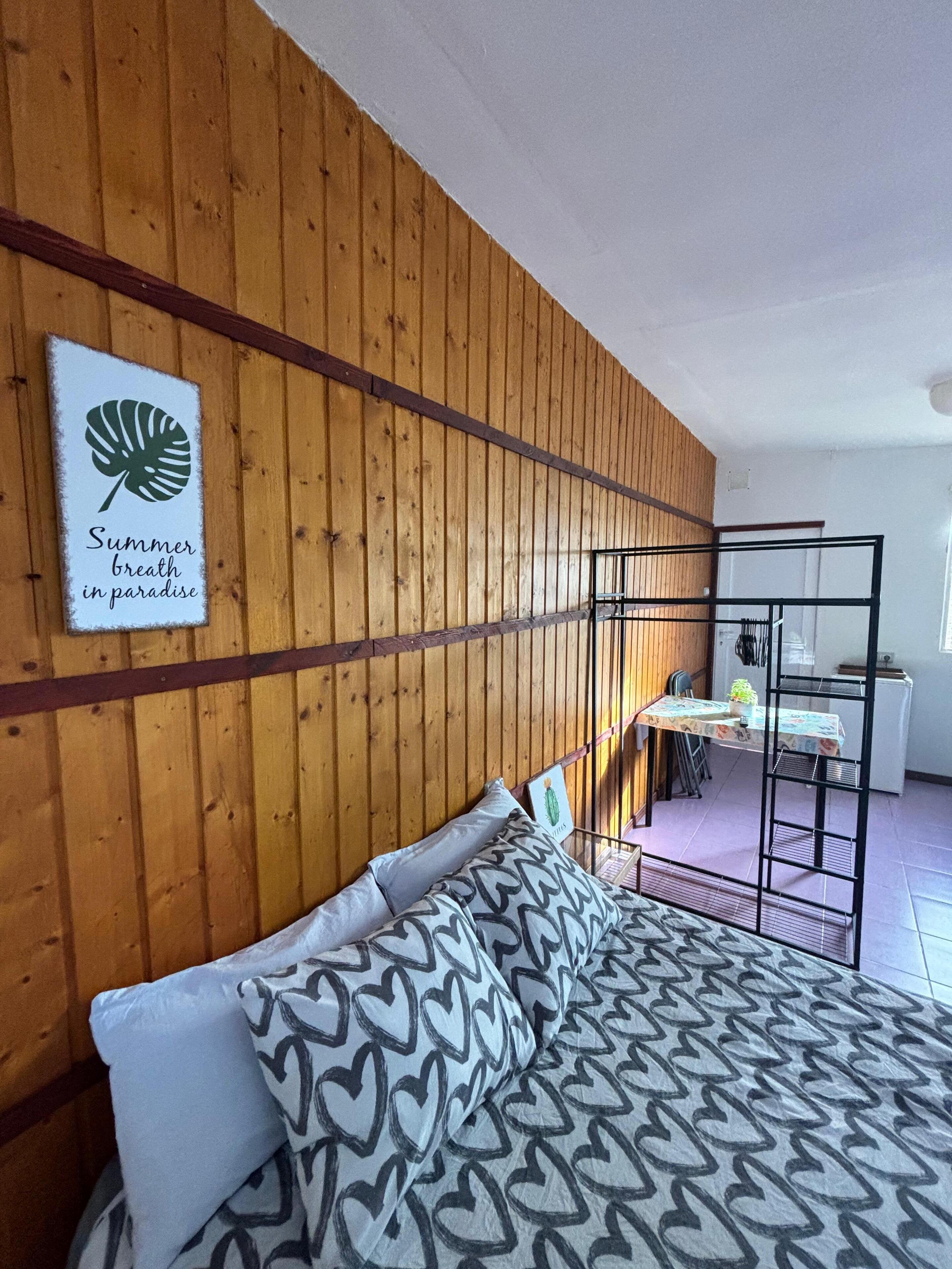 A room with a wooden wall, a bed with patterned pillows, a table with a tablecloth, and a sign with a leaf and text.