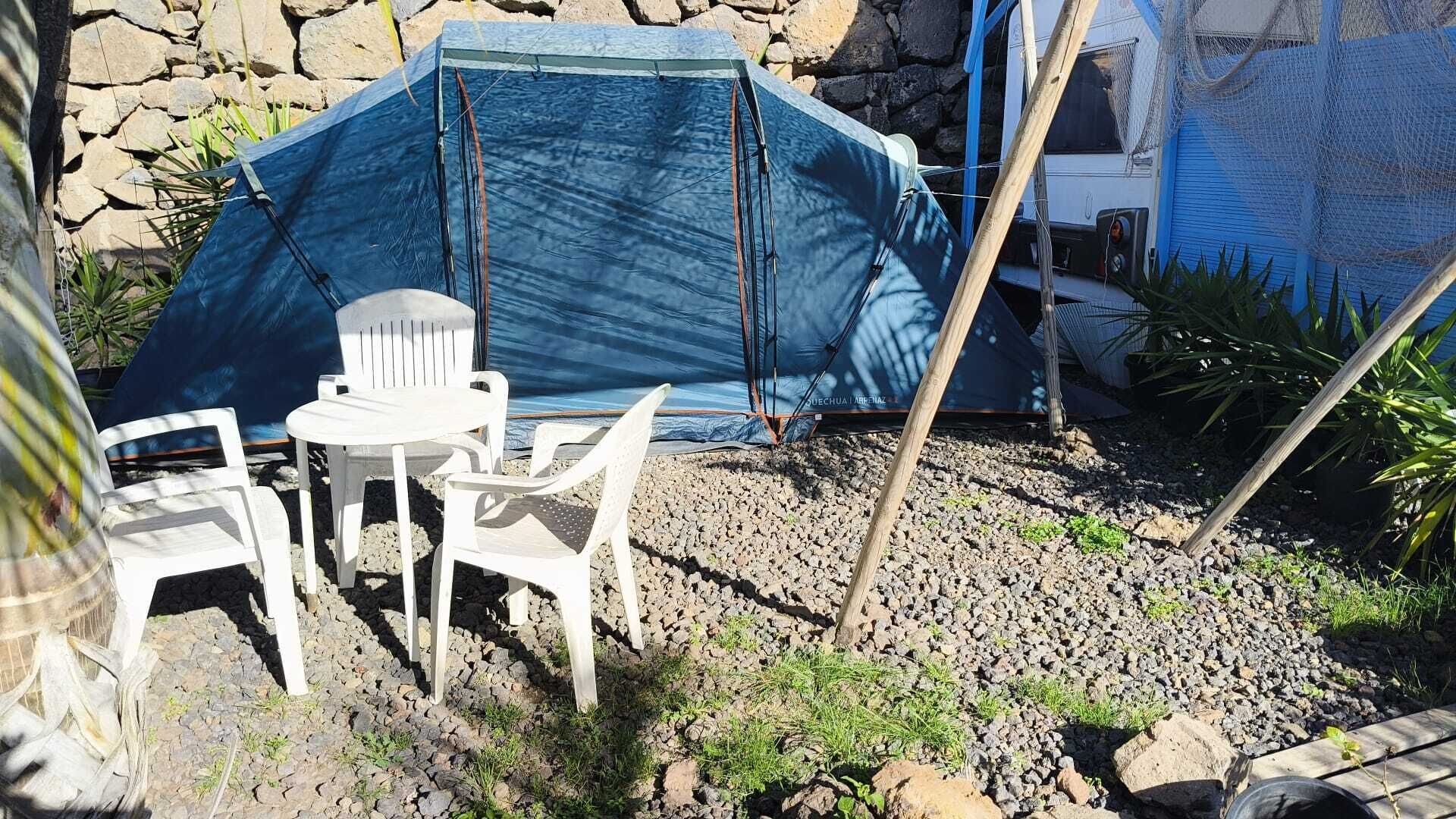 Una tenda con un telo blu, sedie di plastica bianca e un tavolino in una zona rocciosa all'aperto, con un muro di pietra sullo sfondo.