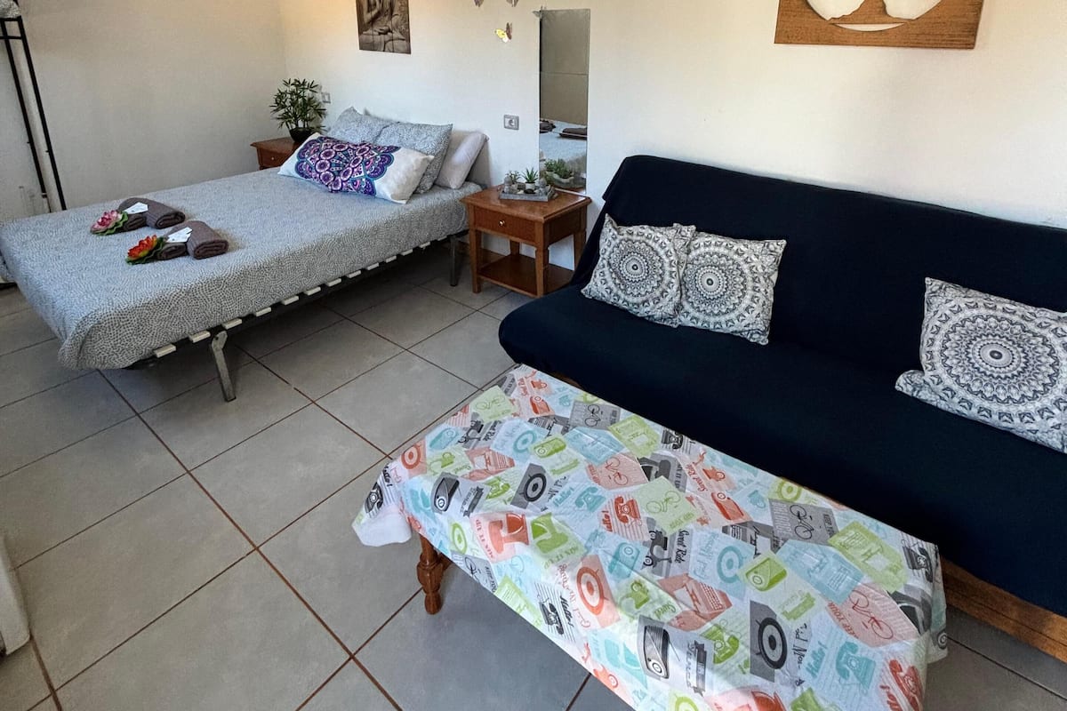 A room with a bed, a sofa, and a small table. There is a wall decoration and a potted plant.
