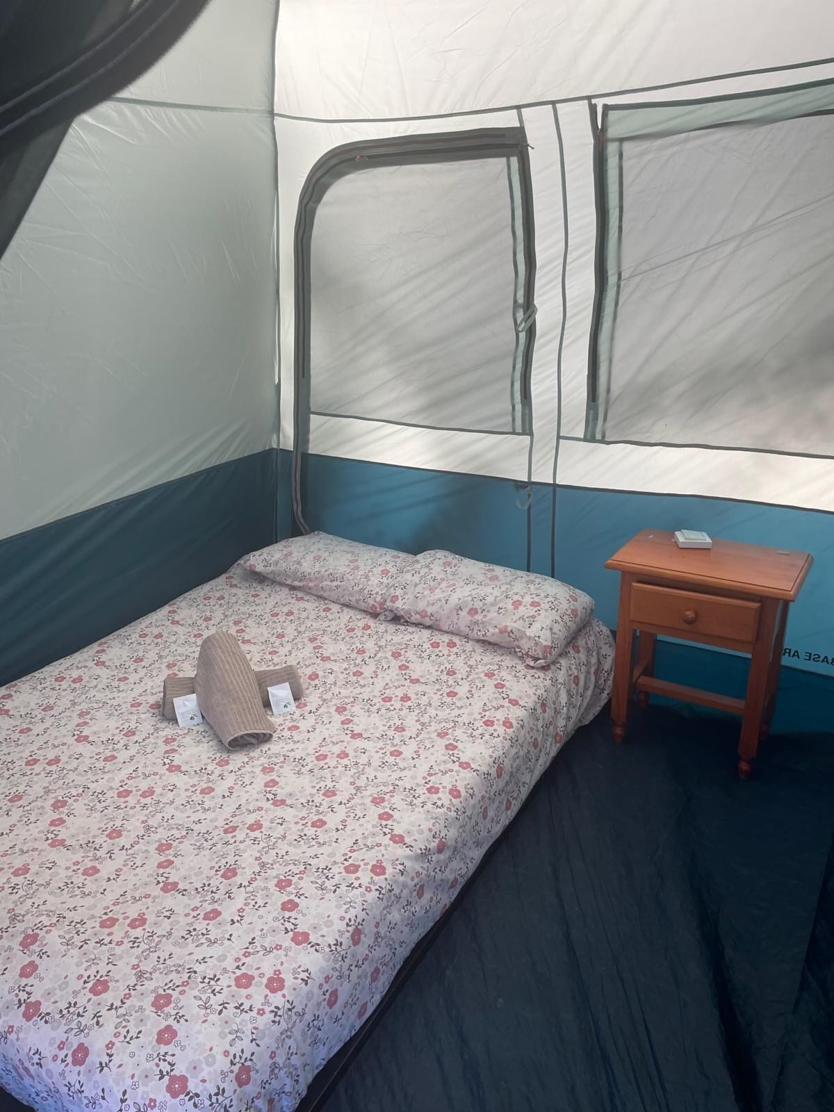 A single bed with a floral bedspread and a pillow, a small wooden nightstand with a lamp, and a tent-like structure with a window.