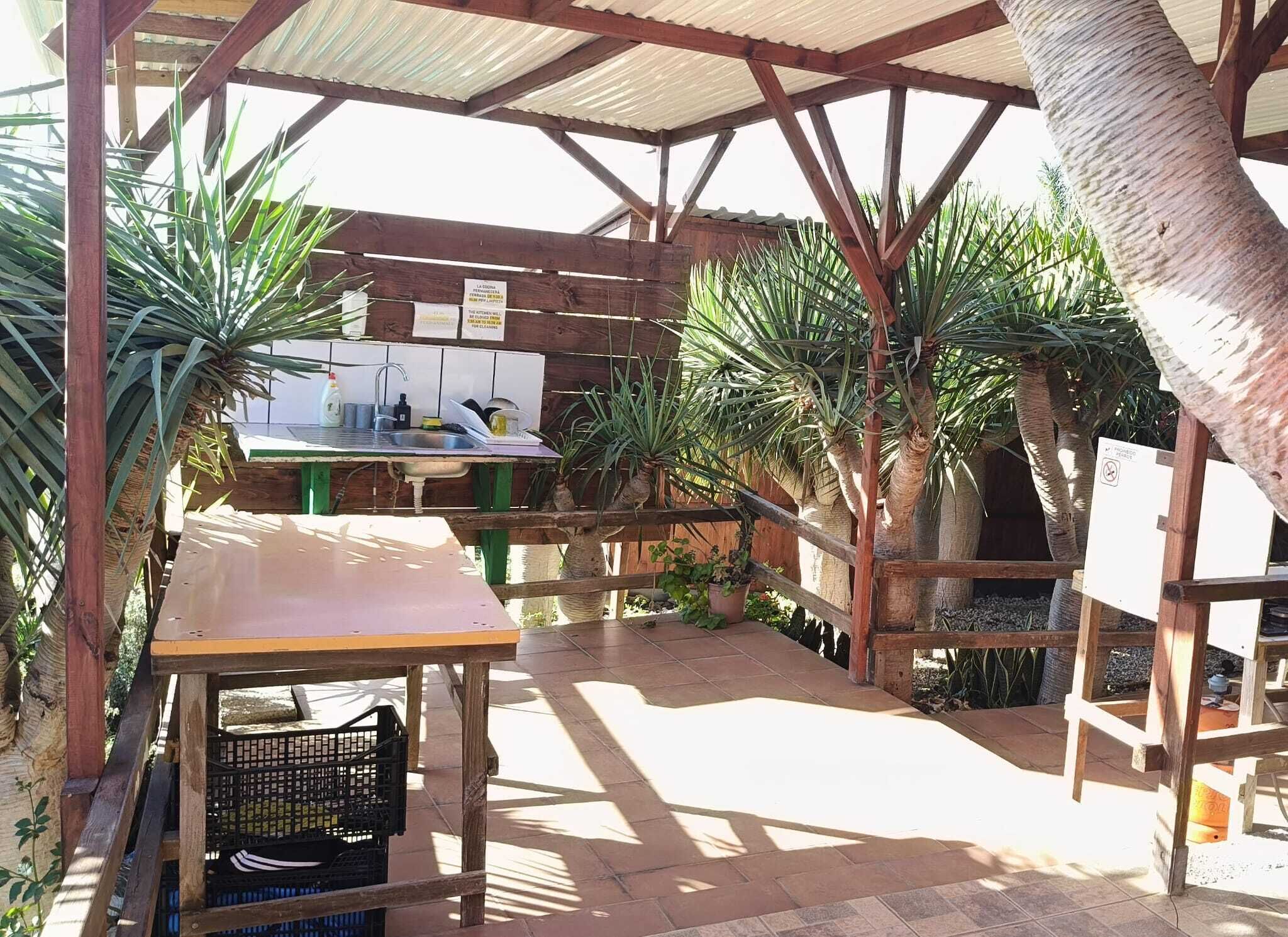 A covered outdoor area with a wooden table and a sink, surrounded by potted plants and a wooden fence.