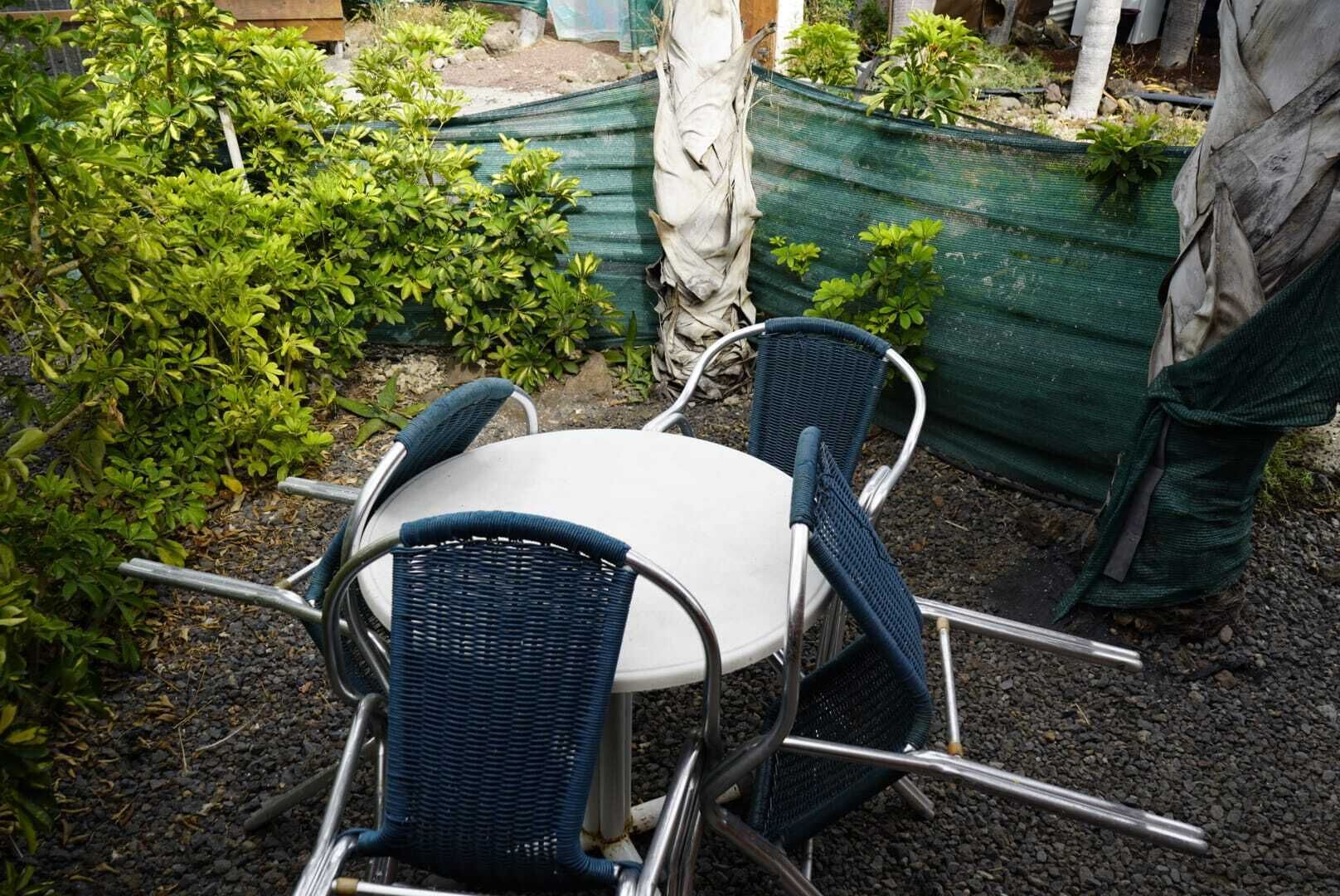 Una mesa redonda con cuatro sillas, ubicada al aire libre sobre un terreno de grava y con follaje de fondo.