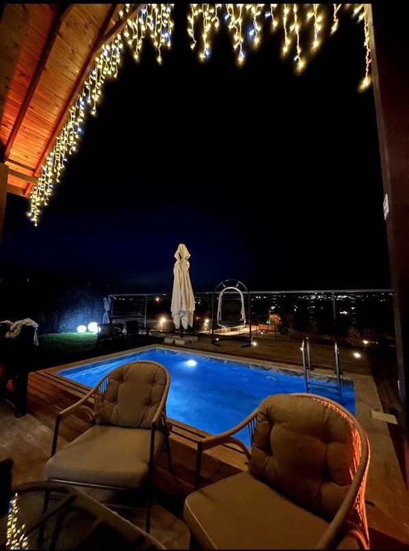 Un área de piscina al aire libre con sillas de descanso, una zona de estar techada y luces colgantes.