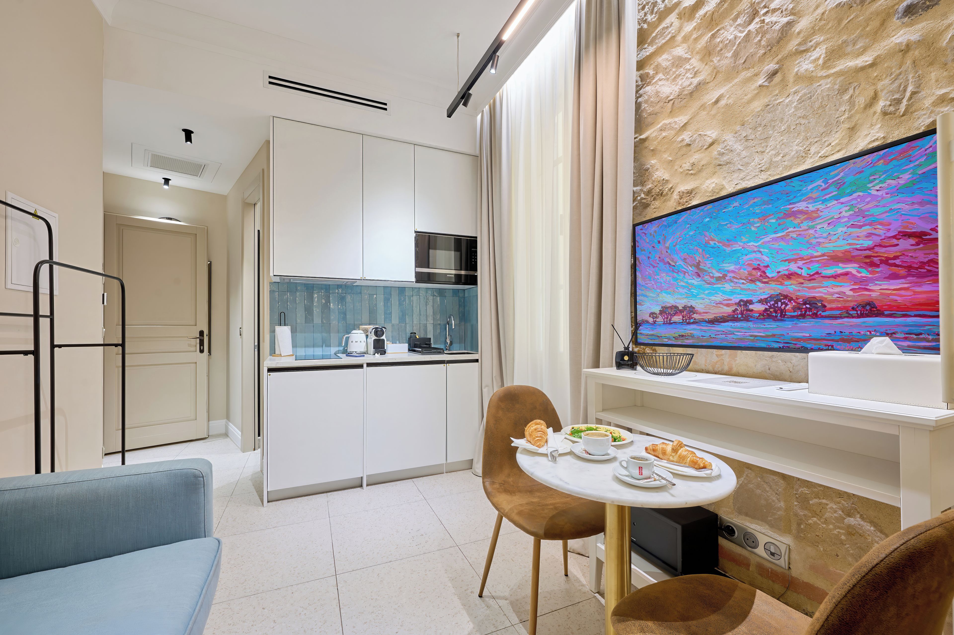 A modern kitchen with white cabinets, a microwave, and a coffee machine. There is a dining area with a table, chairs, and a painting on the wall.
