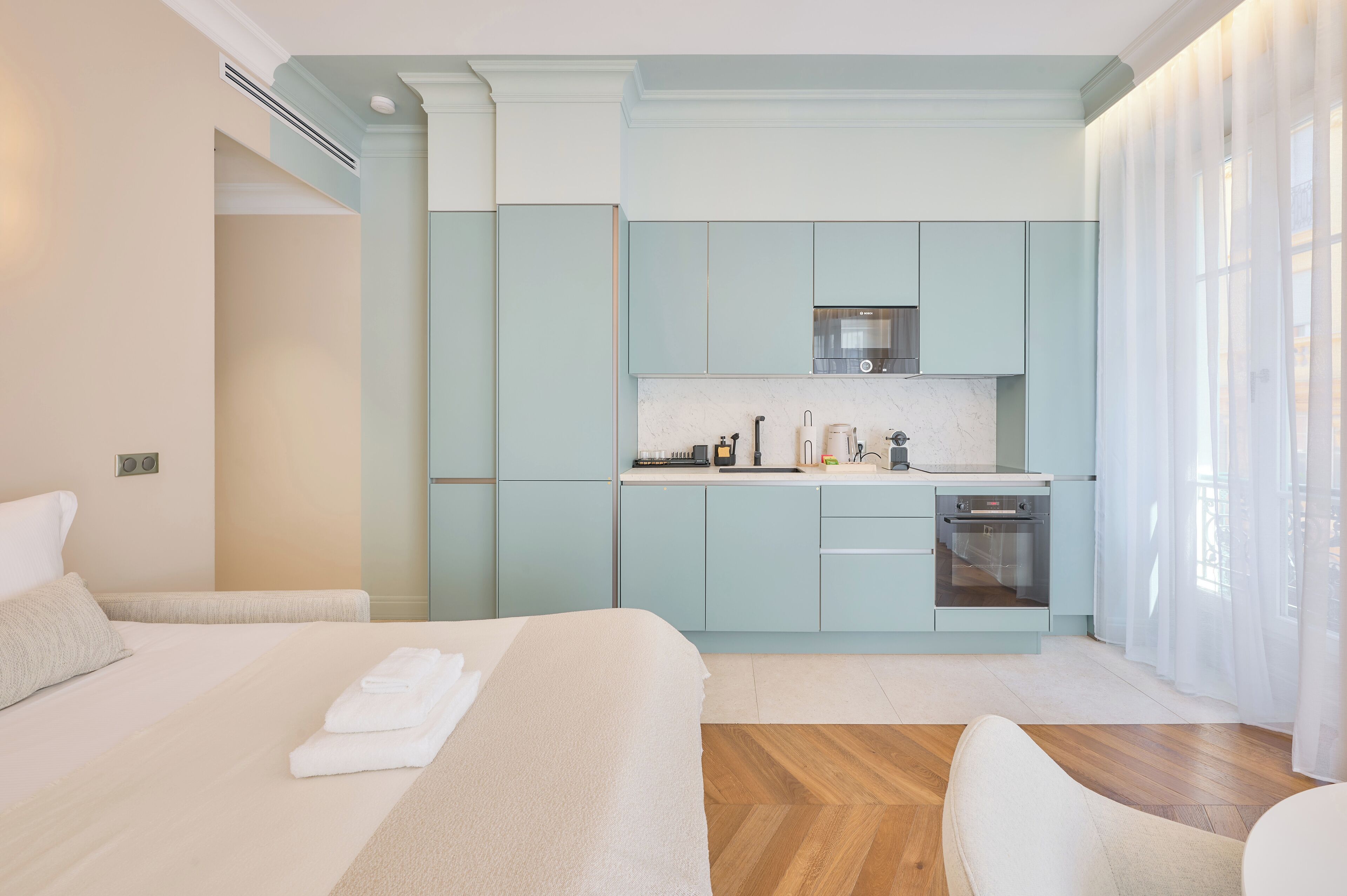 A modern hotel room with a large bed, a separate kitchenette, and a view of the city.