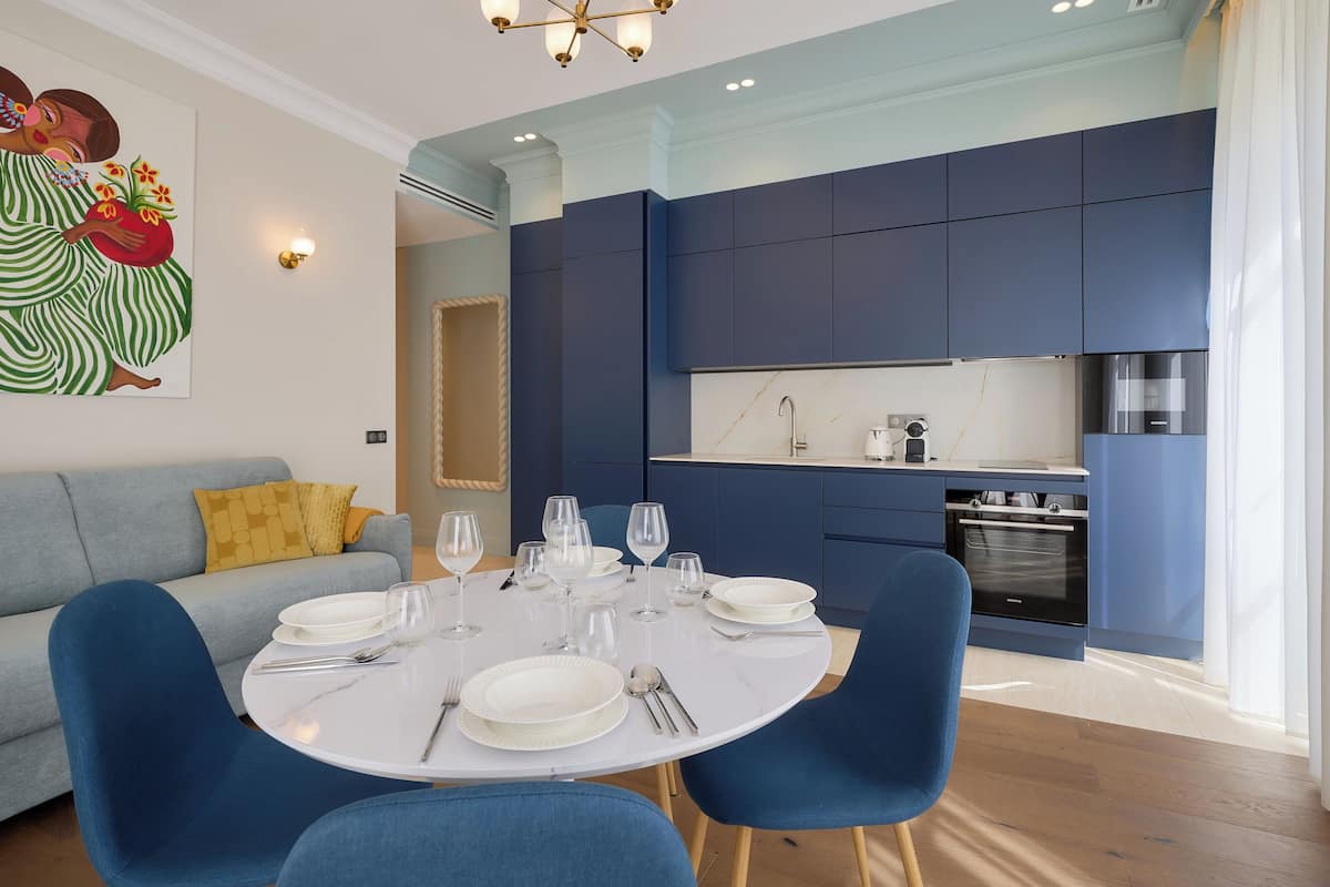 A modern kitchen with blue cabinets, a dining area with a round table set for four, and a grey sofa with a yellow pillow.