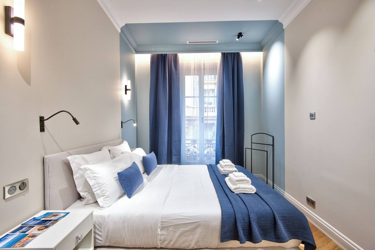 A neatly arranged bedroom with a bed, pillows, a bedside table with a magazine, and a window with curtains.