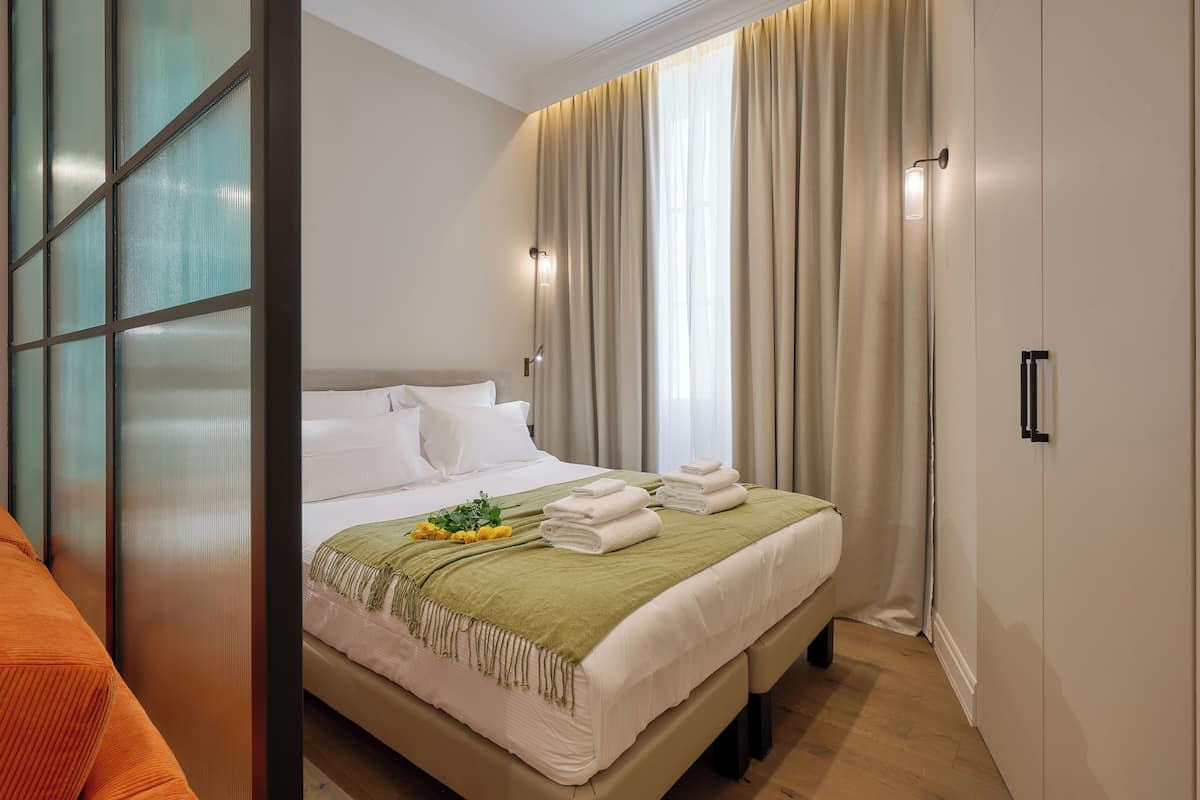 A neatly made bed with white linens and a green throw, a bedside table with a lamp, and a window with curtains.