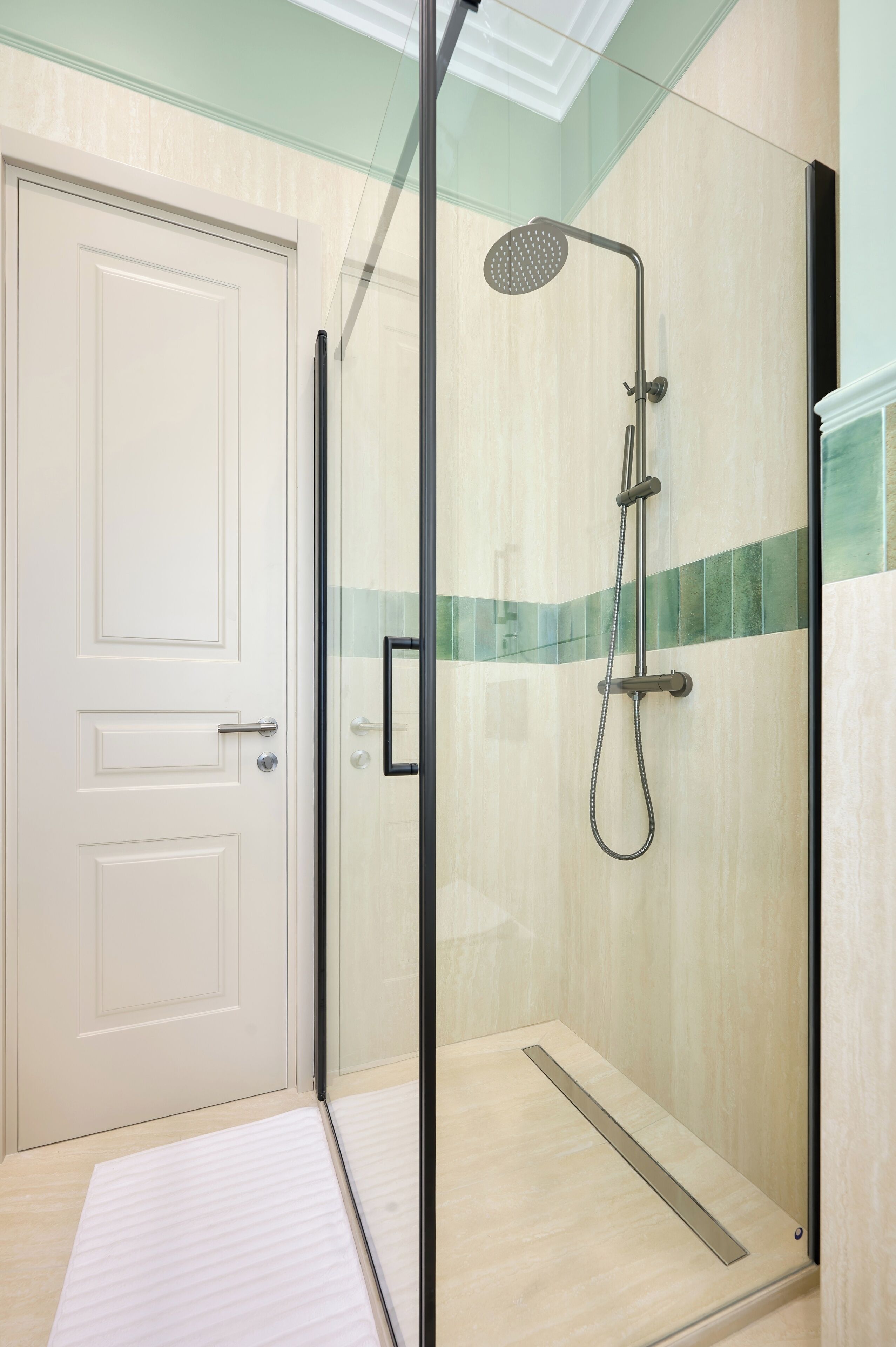 Une douche fermée par des parois en verre, dotée d’un pommeau de douche à main, d’une porte avec une poignée et d’un mur recouvert de carreaux de couleur claire avec une bande verte.