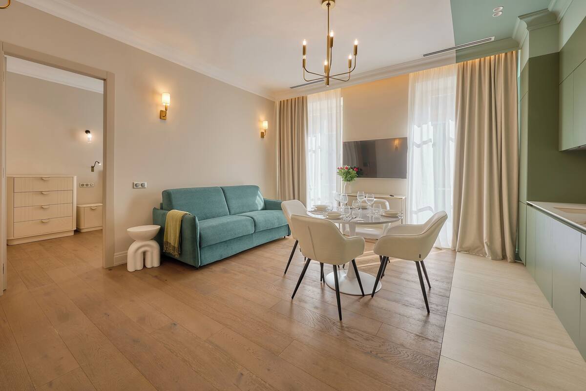 A modern living room with a teal sofa, a dining table with chairs, and a kitchen area with a large window and curtains.